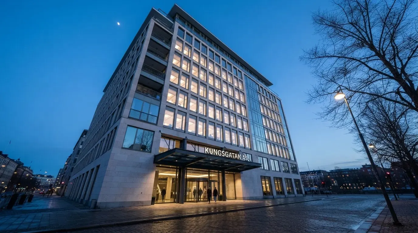 En modern kontorsbyggnad i skymning fotograferad underifrån med tända fönster mot en klarblå himmel i en svensk stad.